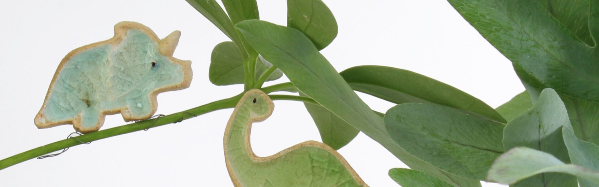 Two ceramic dinosaur figurines perched on a plant. One is a blue triceratops and the other is a green brontosaurus.
