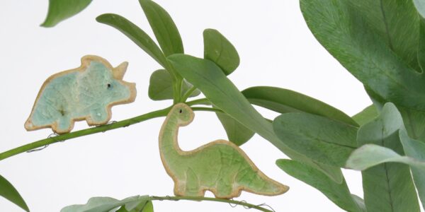 Two ceramic dinosaur figurines perched on a plant. One is a blue triceratops and the other is a green brontosaurus.