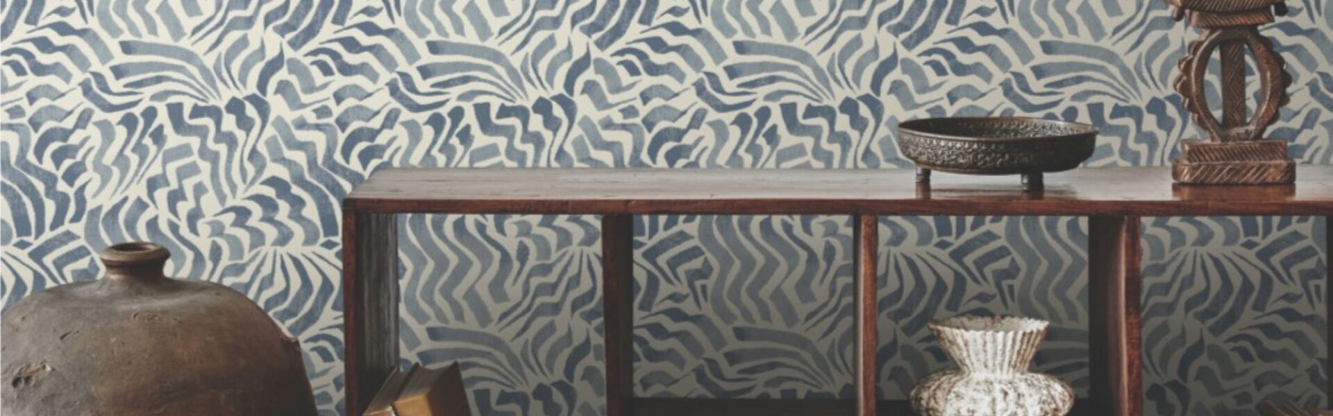 The image shows a room with a feature wall covered in a blue and white patterned wallpaper. The pattern is abstract and wavy, resembling stylised flowers or clouds. The wall is complemented by a wooden console table holding a collection of books, a vase, and a decorative bowl. A large ceramic jar stands to the left of the table, adding to the room's eclectic and cosy atmosphere.