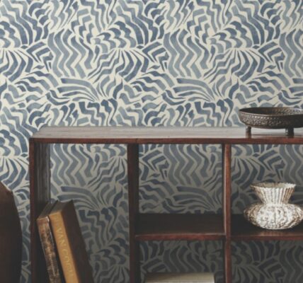 The image shows a room with a feature wall covered in a blue and white patterned wallpaper. The pattern is abstract and wavy, resembling stylised flowers or clouds. The wall is complemented by a wooden console table holding a collection of books, a vase, and a decorative bowl. A large ceramic jar stands to the left of the table, adding to the room's eclectic and cosy atmosphere.
