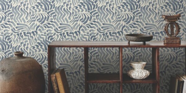 The image shows a room with a feature wall covered in a blue and white patterned wallpaper. The pattern is abstract and wavy, resembling stylised flowers or clouds. The wall is complemented by a wooden console table holding a collection of books, a vase, and a decorative bowl. A large ceramic jar stands to the left of the table, adding to the room's eclectic and cosy atmosphere.
