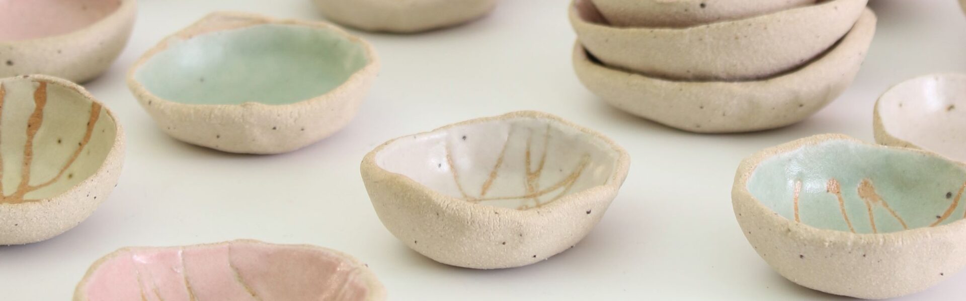 A collection of small, handmade ceramic bowls. They are arranged in groups on a white surface. The bowls vary in size and shape, and are decorated with different colours and patterns. Some bowls are plain white, while others are glazed in pastel colours such as pink, blue, and green. Some bowls have simple designs, such as lines or dots, while others have more intricate patterns.