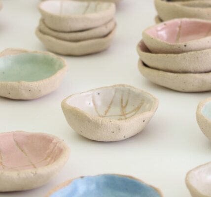 A collection of small, handmade ceramic bowls. They are arranged in groups on a white surface. The bowls vary in size and shape, and are decorated with different colours and patterns. Some bowls are plain white, while others are glazed in pastel colours such as pink, blue, and green. Some bowls have simple designs, such as lines or dots, while others have more intricate patterns.