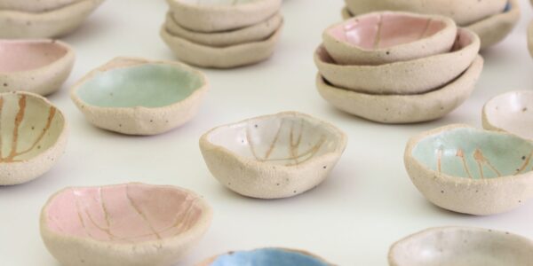 A collection of small, handmade ceramic bowls. They are arranged in groups on a white surface. The bowls vary in size and shape, and are decorated with different colours and patterns. Some bowls are plain white, while others are glazed in pastel colours such as pink, blue, and green. Some bowls have simple designs, such as lines or dots, while others have more intricate patterns.
