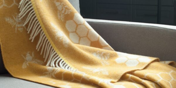 The throw features a distinctive pattern of bees and honeycombs, woven into a soft, mustard yellow wool. The fringes add a touch of texture and style. It seems to be a cozy and visually appealing piece that would complement a variety of home decor styles.
