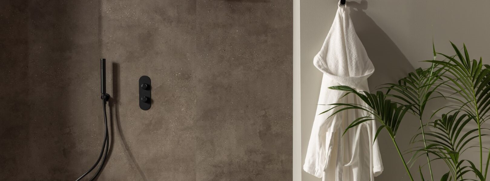 A modern bathroom with dark grey textured panel on the wall. A black rain shower head and matching thermostatic mixer valve are mounted on the wall. A white bathrobe hangs on a hook, and a large palm tree is in a corner.
