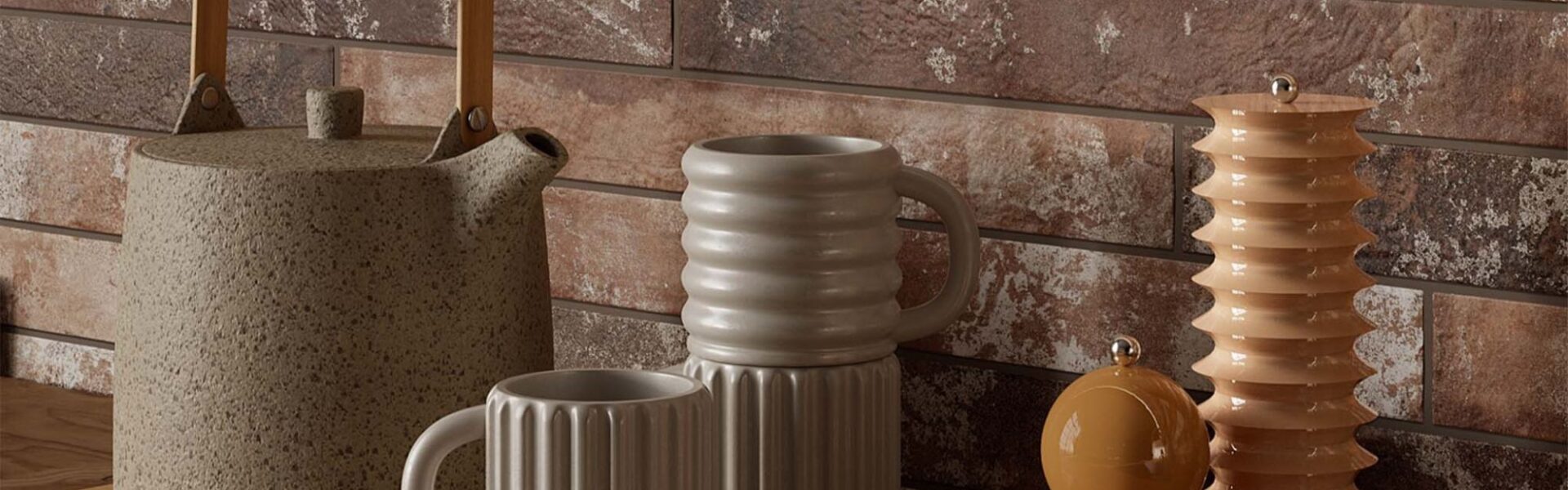 A kitchen countertop with a rustic brick wall backdrop. A wooden tray holds a teapot, a stack of mugs, and a set of salt and pepper shakers. The overall aesthetic is warm and inviting, with a focus on natural materials and earthy tones.