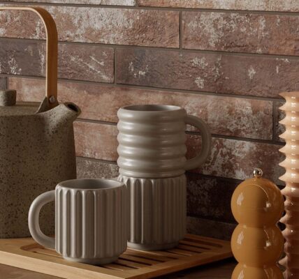 A kitchen countertop with a rustic brick wall backdrop. A wooden tray holds a teapot, a stack of mugs, and a set of salt and pepper shakers. The overall aesthetic is warm and inviting, with a focus on natural materials and earthy tones.