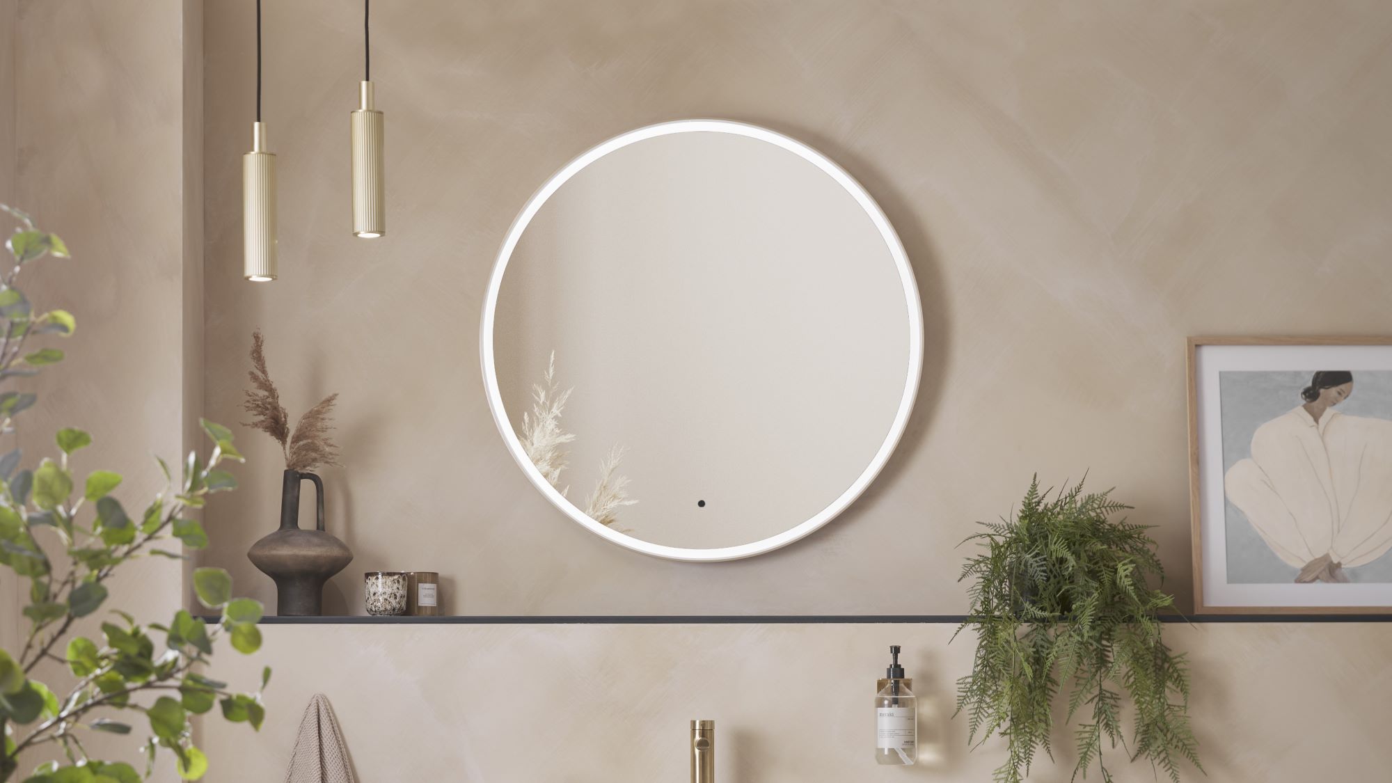 A round, illuminated mirror with a brass frame. It is hanging on a wall in a modern bathroom setting. There are also two gold pendant lights, a vase with dried flowers, a framed picture, and a potted plant.