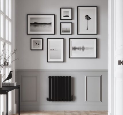 The images shows a single panel horizontal radiator with a matte black finish. It is positioned below a gallery wall of framed photographs. The hallway has grey walls, a wooden floor, and a door leading to another room.