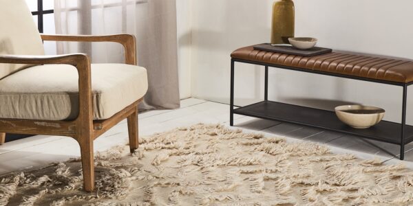 A living room with a beige rug, a wooden armchair, a metal bench with a leather seat, and a vase with dried flowers. The overall style of the room is bohemian and cosy.