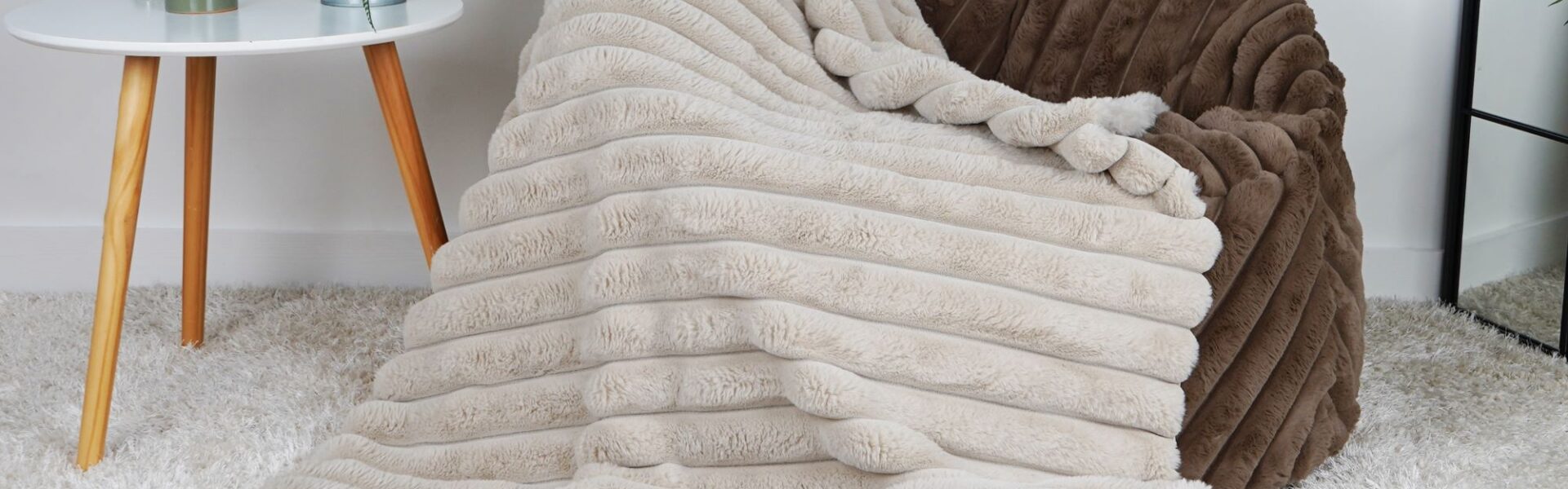 The image shows a faux fur throw blanket draped over a faux fur bean bag chair. The bean bag chair is a light brown colour, and the throw blanket is a light beige colour. The throw blanket has a ribbed texture and is very soft. The bean bag chair is placed in a living room setting, with a white coffee table and a potted plant nearby.