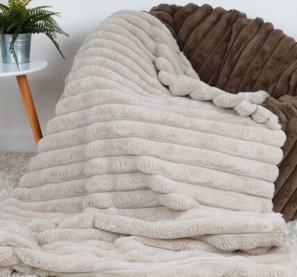 The image shows a faux fur throw blanket draped over a faux fur bean bag chair. The bean bag chair is a light brown colour, and the throw blanket is a light beige colour. The throw blanket has a ribbed texture and is very soft. The bean bag chair is placed in a living room setting, with a white coffee table and a potted plant nearby.