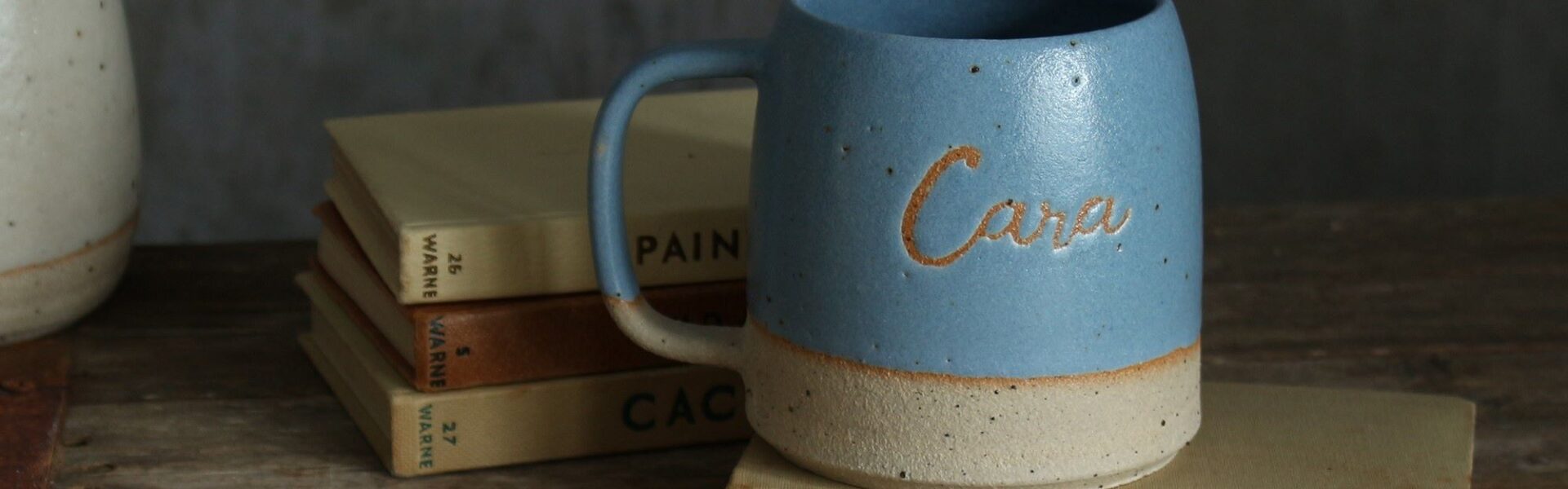 The image conveys a sense of warmth and personalisation. The mug feels like a cherished object with the name "Cara" etched into it, and the books suggest a cosy, personal space.