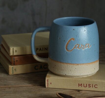 The image conveys a sense of warmth and personalisation. The mug feels like a cherished object with the name "Cara" etched into it, and the books suggest a cosy, personal space.