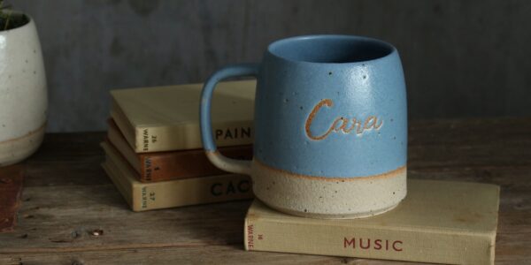 The image conveys a sense of warmth and personalisation. The mug feels like a cherished object with the name "Cara" etched into it, and the books suggest a cosy, personal space.