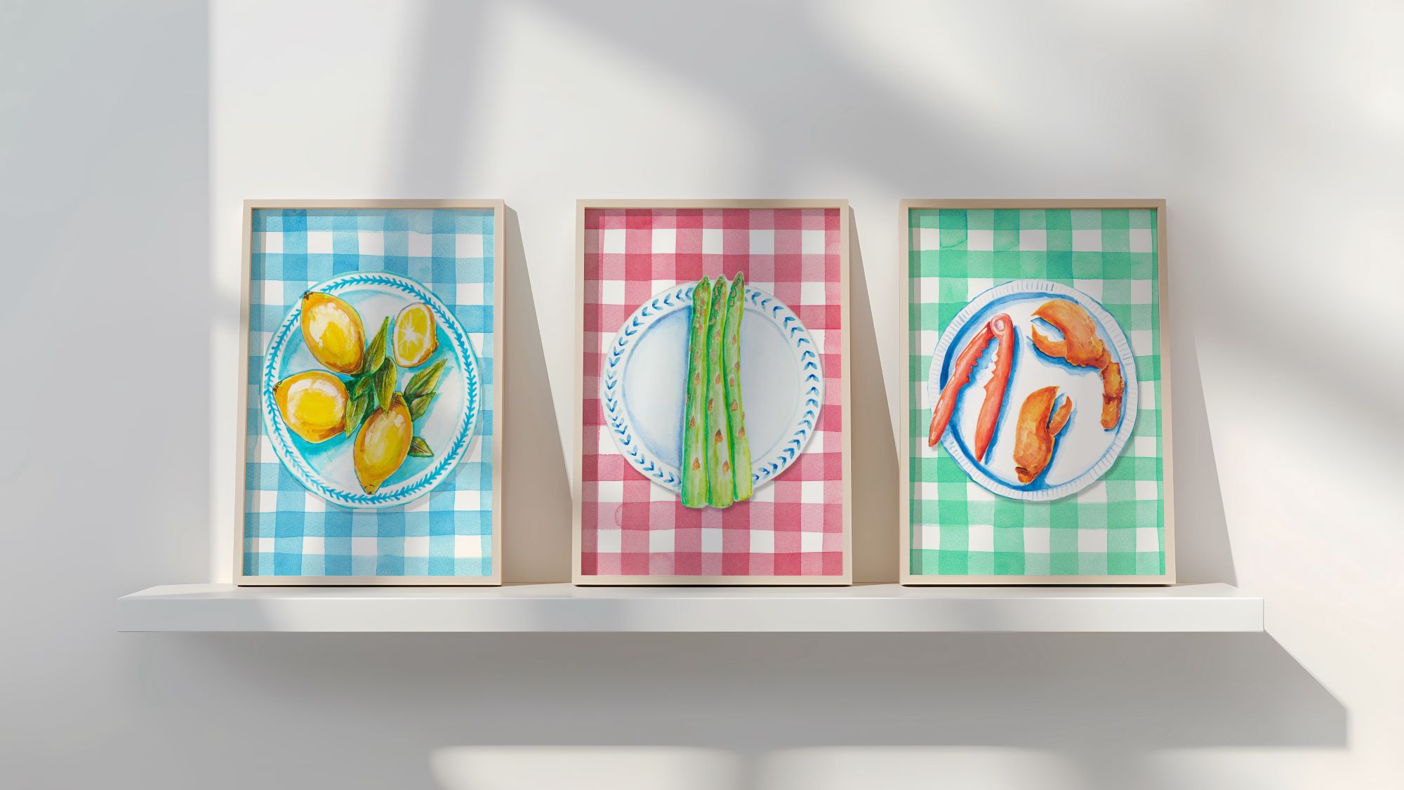 Three framed illustrations of food on plates. The left image shows lemons on a blue and white plate against a blue and white gingham background. The middle image shows asparagus on a blue and white plate against a red and white gingham background. The right image shows lobster claws on a blue and white plate against a green and white gingham background.