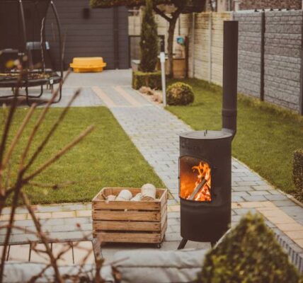 A cosy outdoor space featuring a modern black wood-burning stove with a visible flame, set on a paved patio surrounded by a neatly manicured garden. A wooden box filled with logs sits nearby, creating a warm and inviting atmosphere.