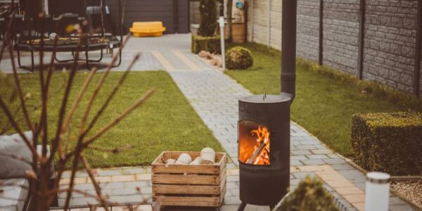A cosy outdoor space featuring a modern black wood-burning stove with a visible flame, set on a paved patio surrounded by a neatly manicured garden. A wooden box filled with logs sits nearby, creating a warm and inviting atmosphere.
