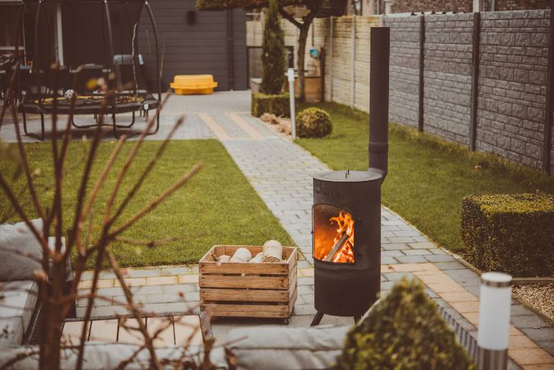 A cosy outdoor space featuring a modern black wood-burning stove with a visible flame, set on a paved patio surrounded by a neatly manicured garden. A wooden box filled with logs sits nearby, creating a warm and inviting atmosphere.