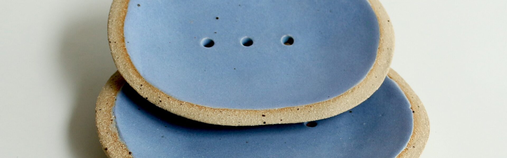 Two blue ceramic soap dishes with a raised rim and drainage holes. The soap dishes are slightly textured and have a natural, organic look.