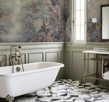 A spacious bathroom with a freestanding white clawfoot bathtub, a patterned geometric tile floor, and a wallpapered wall featuring a lush landscape scene. A vanity with a marble sink and a mirror complete the room.