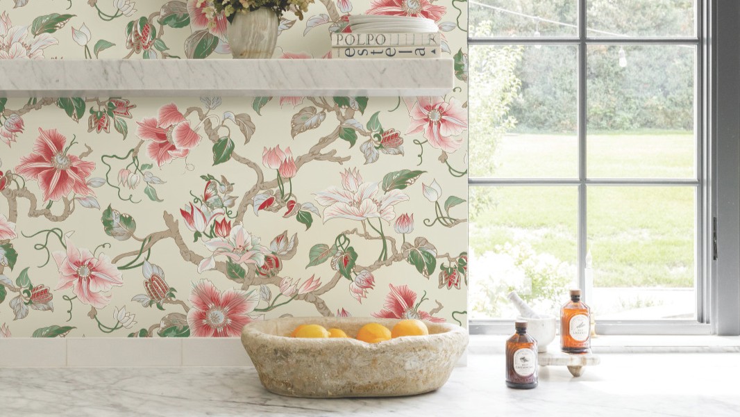 A bright kitchen featuring Ronald Redding Classics Marguerite Vine wallpaper in a pink and green botanical pattern, white marble countertops, a vintage stone bowl filled with oranges, and two bottles on the counter.