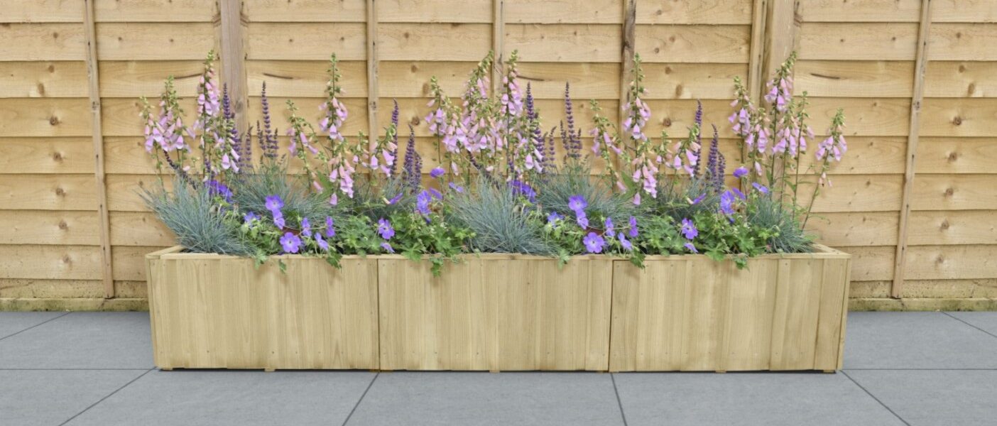A rectangular wooden planter box sits on a grey tiled surface against a light wood fence. The planter is filled with a variety of purple and lavender flowers.