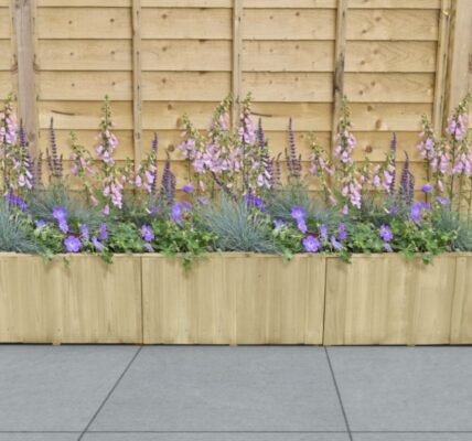A rectangular wooden planter box sits on a grey tiled surface against a light wood fence. The planter is filled with a variety of purple and lavender flowers.