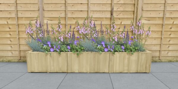 A rectangular wooden planter box sits on a grey tiled surface against a light wood fence. The planter is filled with a variety of purple and lavender flowers.