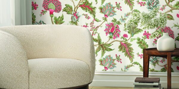 A close-up shot of a cream-coloured, rounded boucle fabric chair with a dark wood base, positioned in front of a wall with a vibrant floral wallpaper featuring pink, green, and white flowers and leaves.