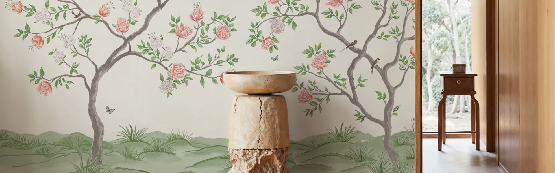 A bright, indoor space featuring a mural-style wallpaper of flowering trees against a light background. A tall, rustic pedestal made of wood and stone holds a simple bowl in the foreground. The room has light wood floors and a doorway leading to an outdoor space with lush greenery.