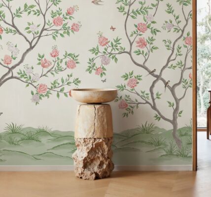 A bright, indoor space featuring a mural-style wallpaper of flowering trees against a light background. A tall, rustic pedestal made of wood and stone holds a simple bowl in the foreground. The room has light wood floors and a doorway leading to an outdoor space with lush greenery.