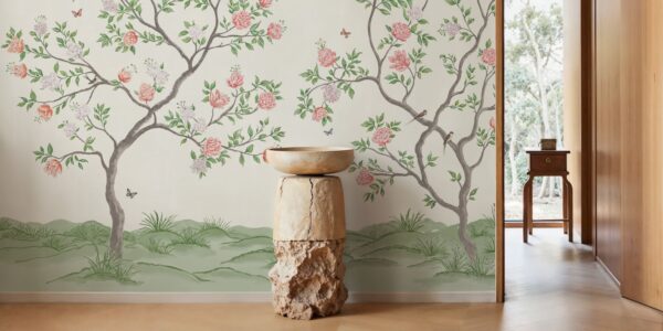 A bright, indoor space featuring a mural-style wallpaper of flowering trees against a light background. A tall, rustic pedestal made of wood and stone holds a simple bowl in the foreground. The room has light wood floors and a doorway leading to an outdoor space with lush greenery.