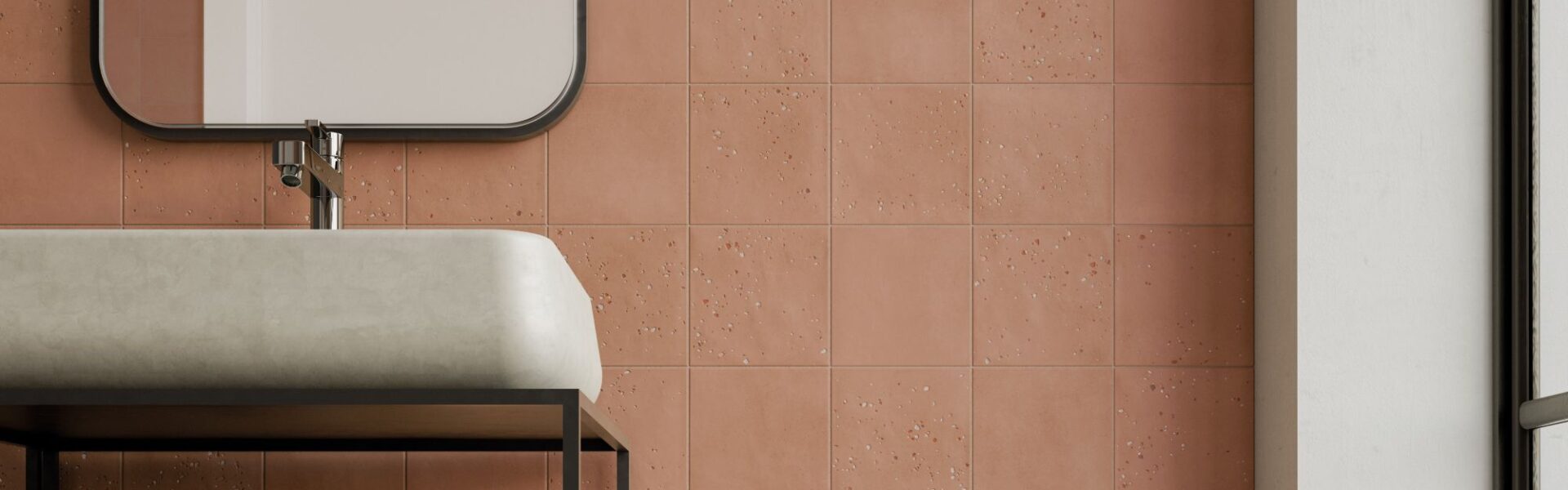 A bathroom wall tiled with square, textured terracotta tiles in a grid pattern. A white sink and black framed mirror are partially visible.