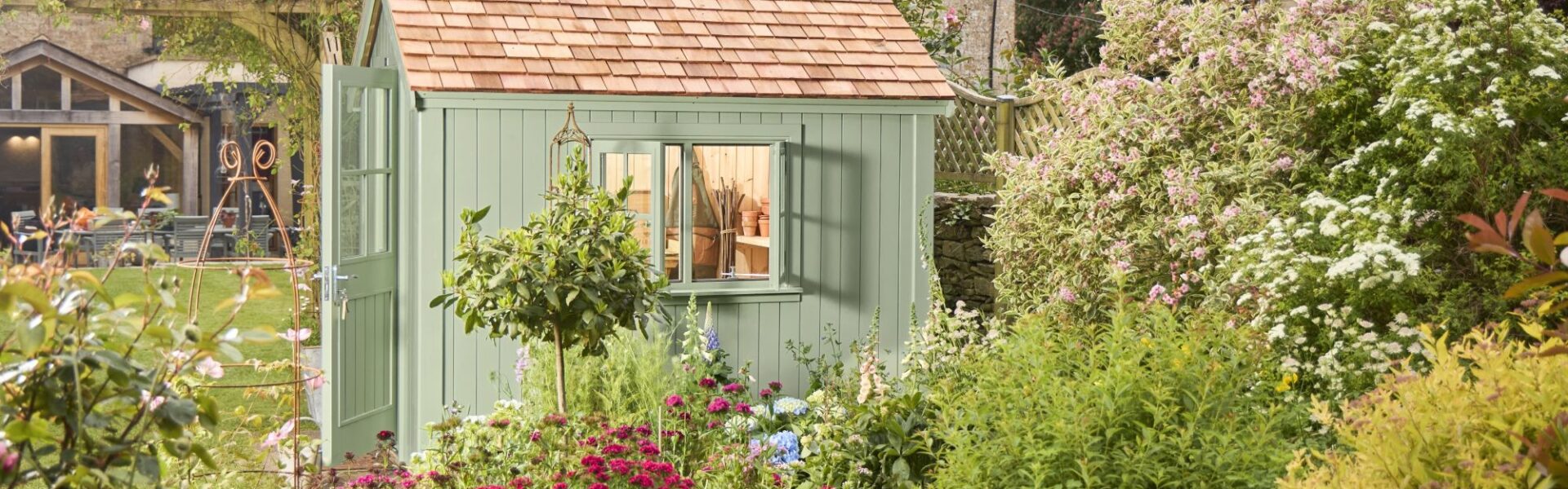 The primary focus is the potting shed, painted in a soft, muted green. It has a traditional design with vertical plank siding and a pitched roof covered in small, rectangular cedar shingles. The double doors have simple panelled detailing.