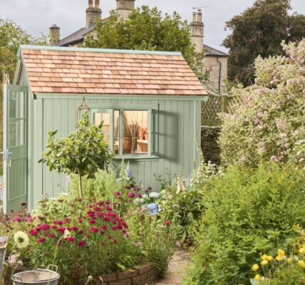 The primary focus is the potting shed, painted in a soft, muted green. It has a traditional design with vertical plank siding and a pitched roof covered in small, rectangular cedar shingles. The double doors have simple panelled detailing.