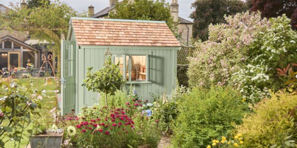 The primary focus is the potting shed, painted in a soft, muted green. It has a traditional design with vertical plank siding and a pitched roof covered in small, rectangular cedar shingles. The double doors have simple panelled detailing.
