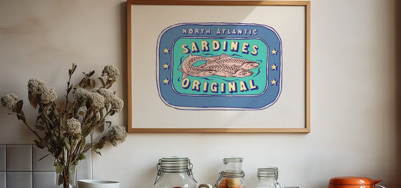 A framed illustration of a vintage sardine can label hangs on a white wall above a kitchen counter. The label features three sardines swimming in a blue oval surrounded by a light blue border.