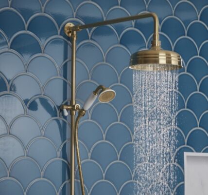 A close-up shot of a brass-finished shower system against a backdrop of blue, fish-scale patterned tiles. Water streams from the overhead rain shower head. A handheld shower wand is also visible on the left.