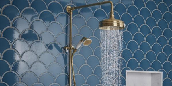 A close-up shot of a brass-finished shower system against a backdrop of blue, fish-scale patterned tiles. Water streams from the overhead rain shower head. A handheld shower wand is also visible on the left.