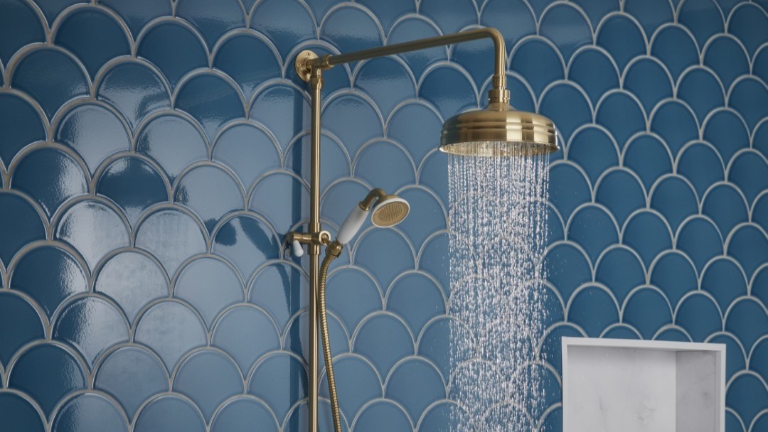 A close-up shot of a brass-finished shower system against a backdrop of blue, fish-scale patterned tiles. Water streams from the overhead rain shower head. A handheld shower wand is also visible on the left.