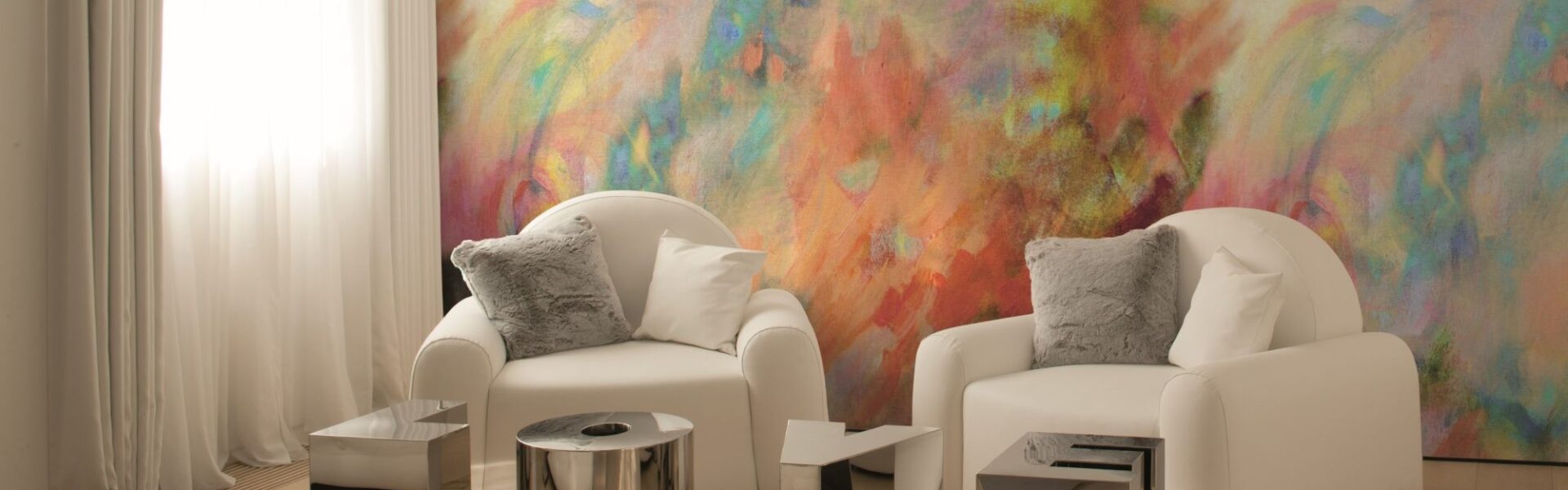 A bright, modern room features two white armchairs facing each other, with a vibrant, abstract painting-like mural on the wall behind them. The mural is a mix of swirling colours including yellow, orange, red, purple, blue, and green.
