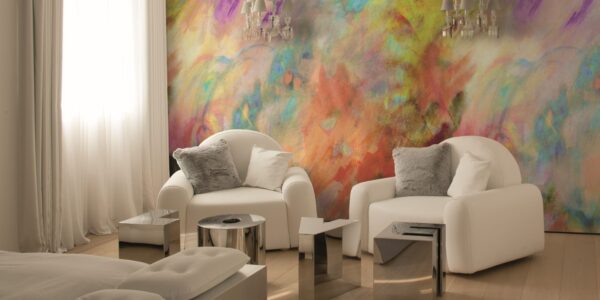 A bright, modern room features two white armchairs facing each other, with a vibrant, abstract painting-like mural on the wall behind them. The mural is a mix of swirling colours including yellow, orange, red, purple, blue, and green.