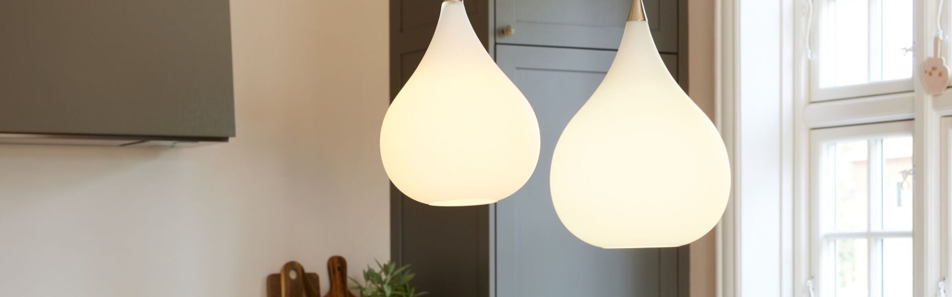 A close-up interior shot shows two teardrop-shaped pendant lights hanging in a kitchen. The lights are made of white, frosted glass and emit a soft, warm glow. They hang from thin black cords and have a small metallic accent at the top.