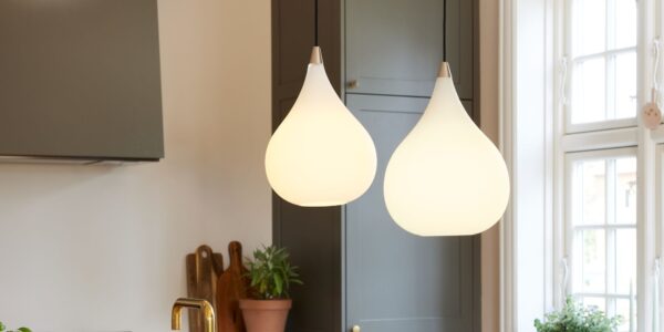 A close-up interior shot shows two teardrop-shaped pendant lights hanging in a kitchen. The lights are made of white, frosted glass and emit a soft, warm glow. They hang from thin black cords and have a small metallic accent at the top.