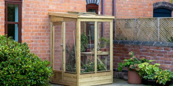 Outdoor image of a wooden lean-to greenhouse with a single door and clear panels, positioned against a brick wall with arched windows. Potted plants are visible inside.