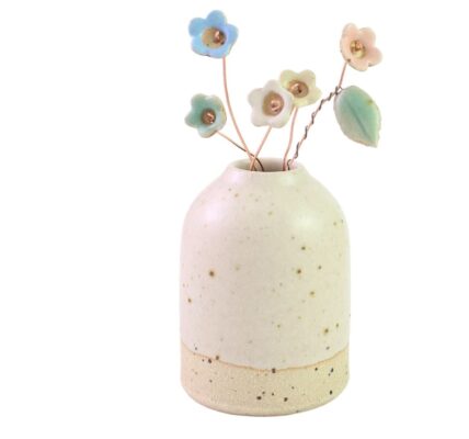 A small, cream-colored ceramic vase with speckled detailing, holding several delicate, pastel-coloured ceramic wildflower stems. The flowers are in shades of blue, pink, and yellow, and are attached to thin, brown wire stems. One stem also features a small, green ceramic leaf.