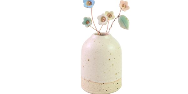 A small, cream-colored ceramic vase with speckled detailing, holding several delicate, pastel-coloured ceramic wildflower stems. The flowers are in shades of blue, pink, and yellow, and are attached to thin, brown wire stems. One stem also features a small, green ceramic leaf.