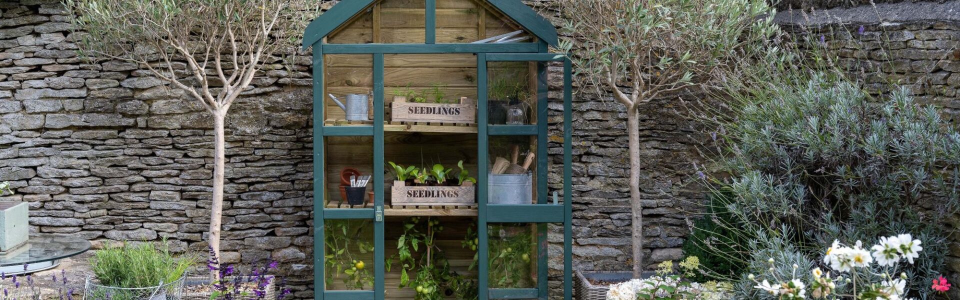 A dark green, A-frame greenhouse with wooden construction and glass panels sits in a garden setting.